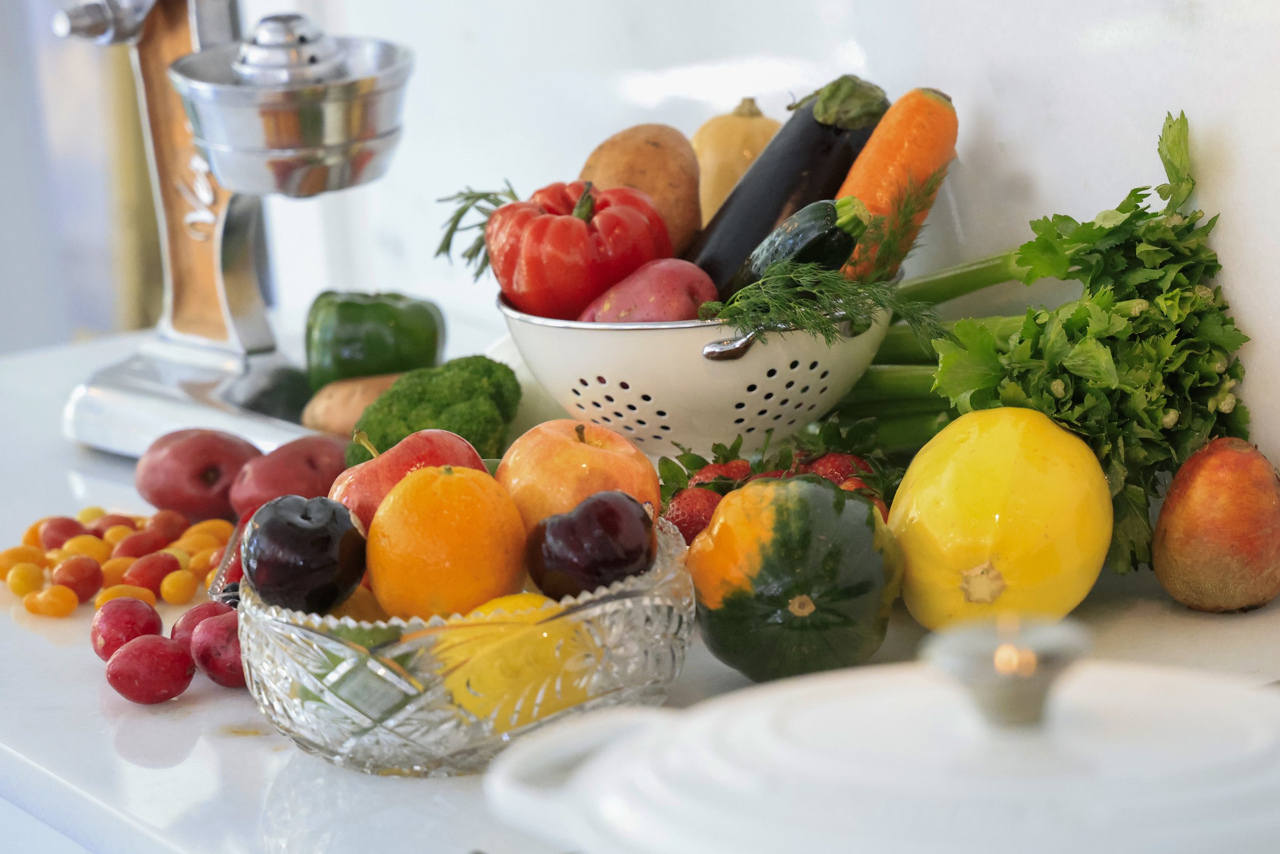 A variety of fresh fruits and vegetables, including tomatoes, apples, oranges, cucumbers, carrots, and broccoli, displayed on a kitchen countertop alongside a vintage juicer and a colander.