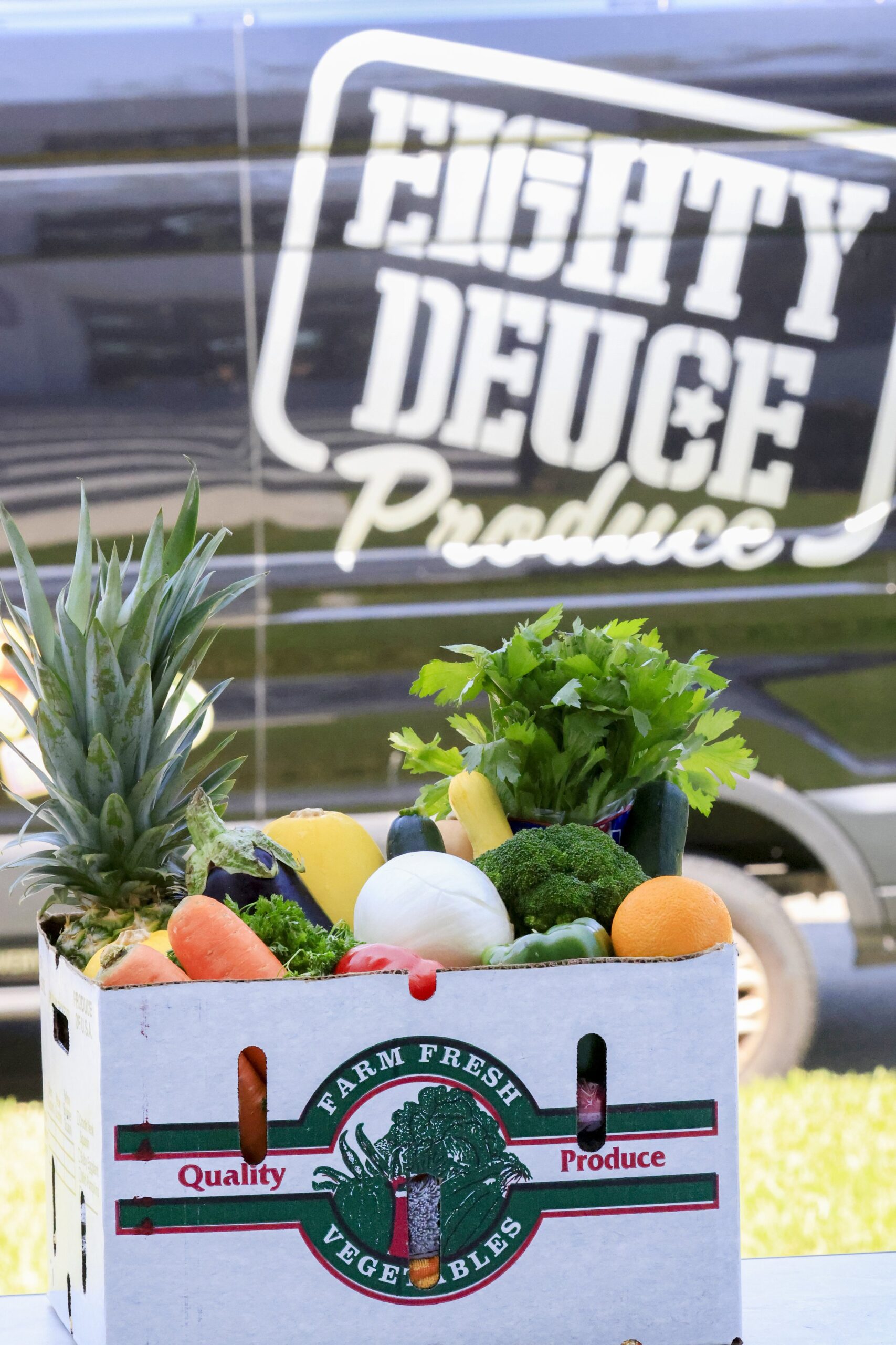 Fresh produce box filled with a variety of vegetables and fruits, including broccoli, carrots, celery, pineapple, and squash, with an "Eighty Deuce Produce" delivery van in the background. Ideal for promoting healthy eating and local farming.