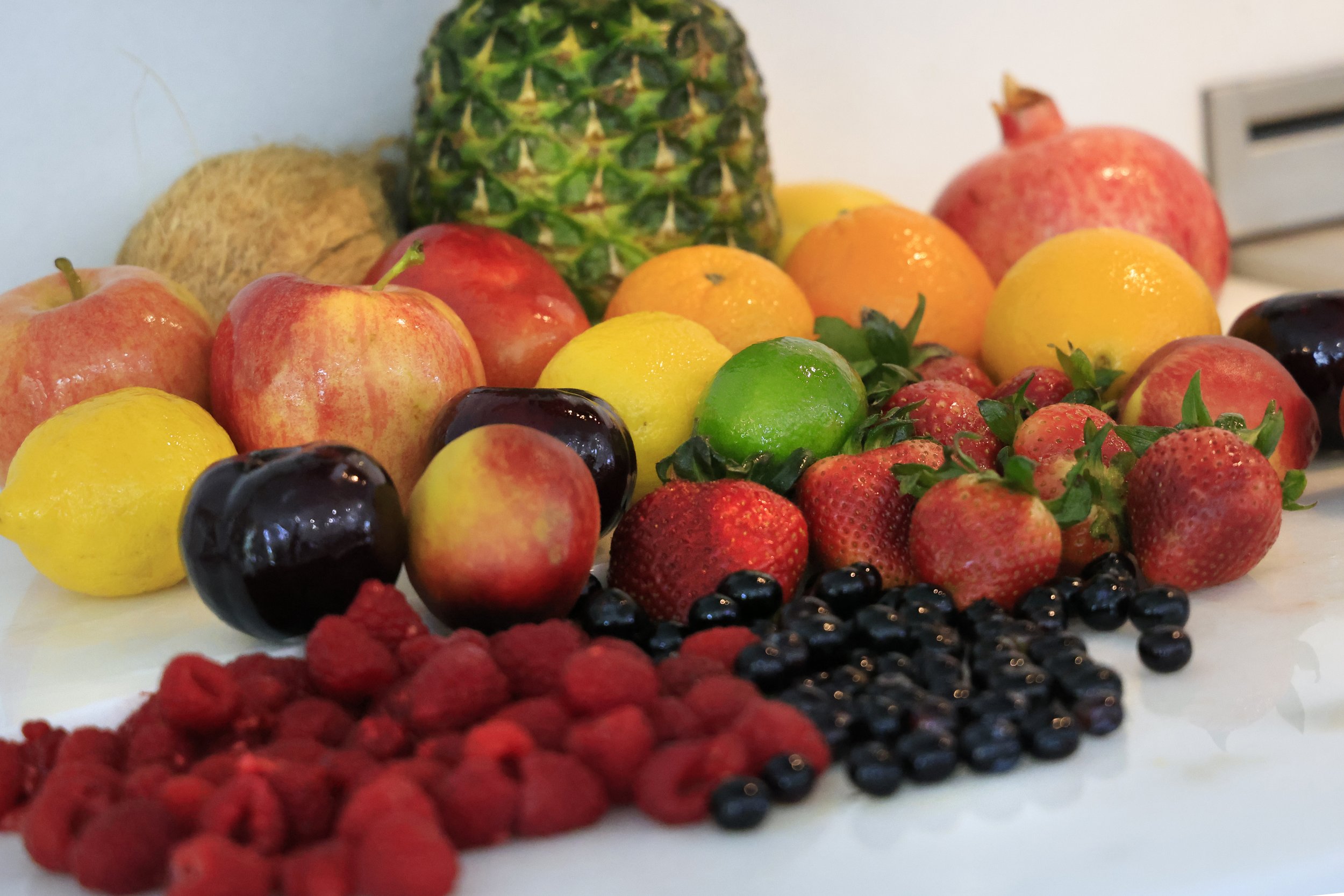 Assorted fresh fruits including apples, lemons, strawberries, blueberries, raspberries, pomegranates, oranges, and a pineapple arranged on a white surface, showcasing vibrant colors and textures.