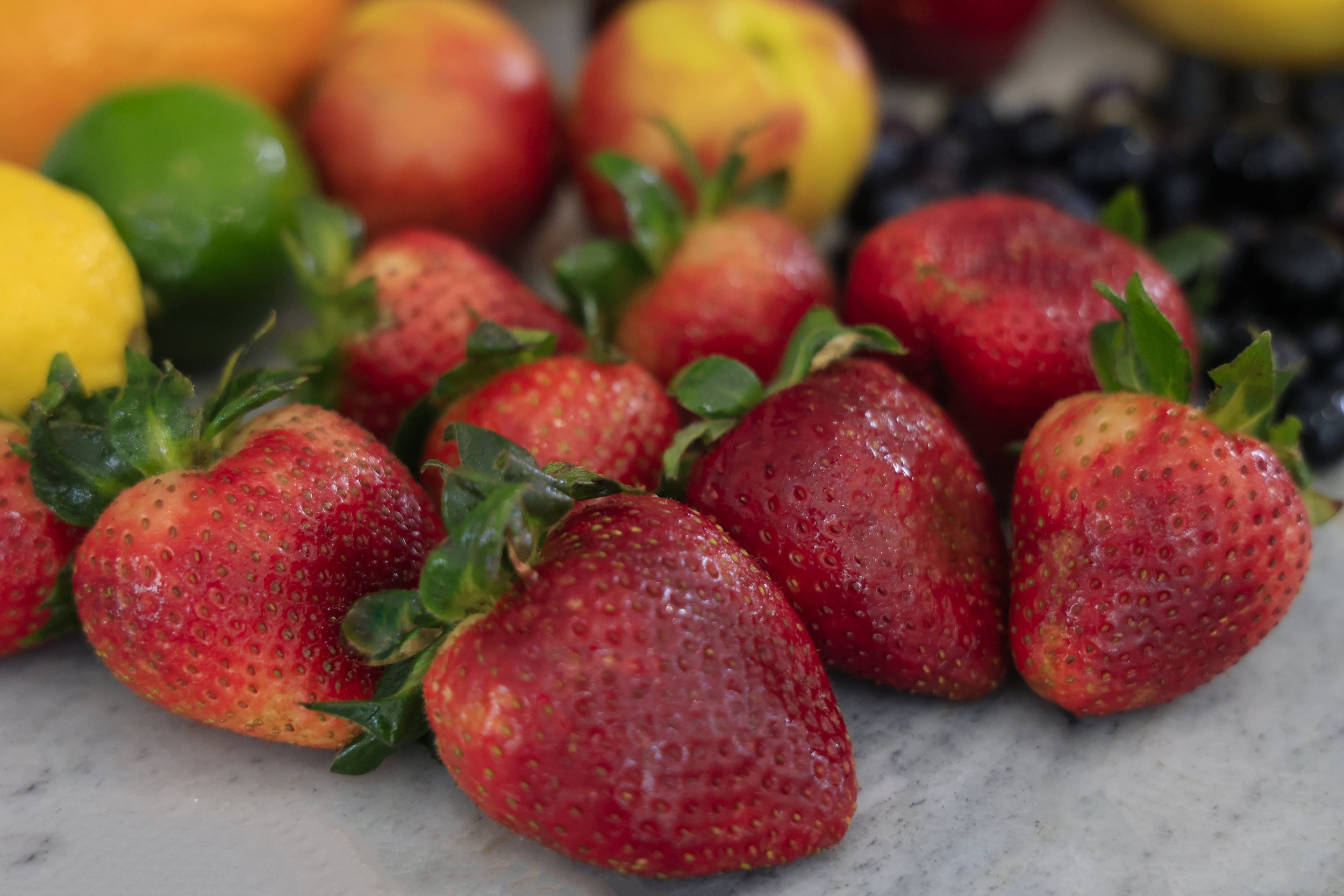 Fresh, ripe strawberries with vibrant red color and green leaves, surrounded by various fruits including lemons, limes, and apples, on a marble surface. Ideal for healthy recipes and summer snacks.