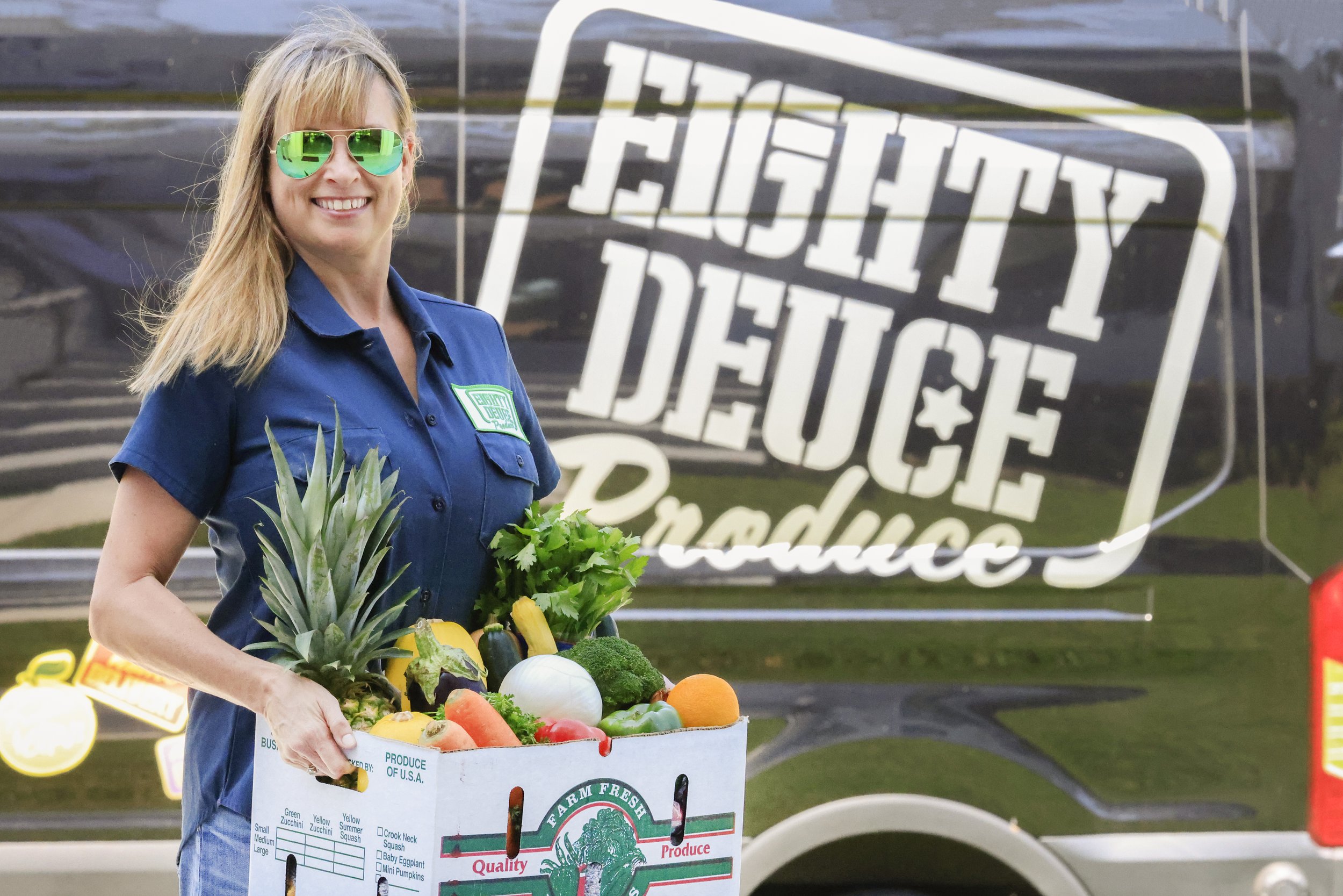 Woman holding a box of fresh produce, including a pineapple, peppers, and broccoli, in front of a vehicle with the "Eighty Deuce Produce" logo. The woman is wearing sunglasses and a blue shirt with a green badge, promoting farm-fresh quality.