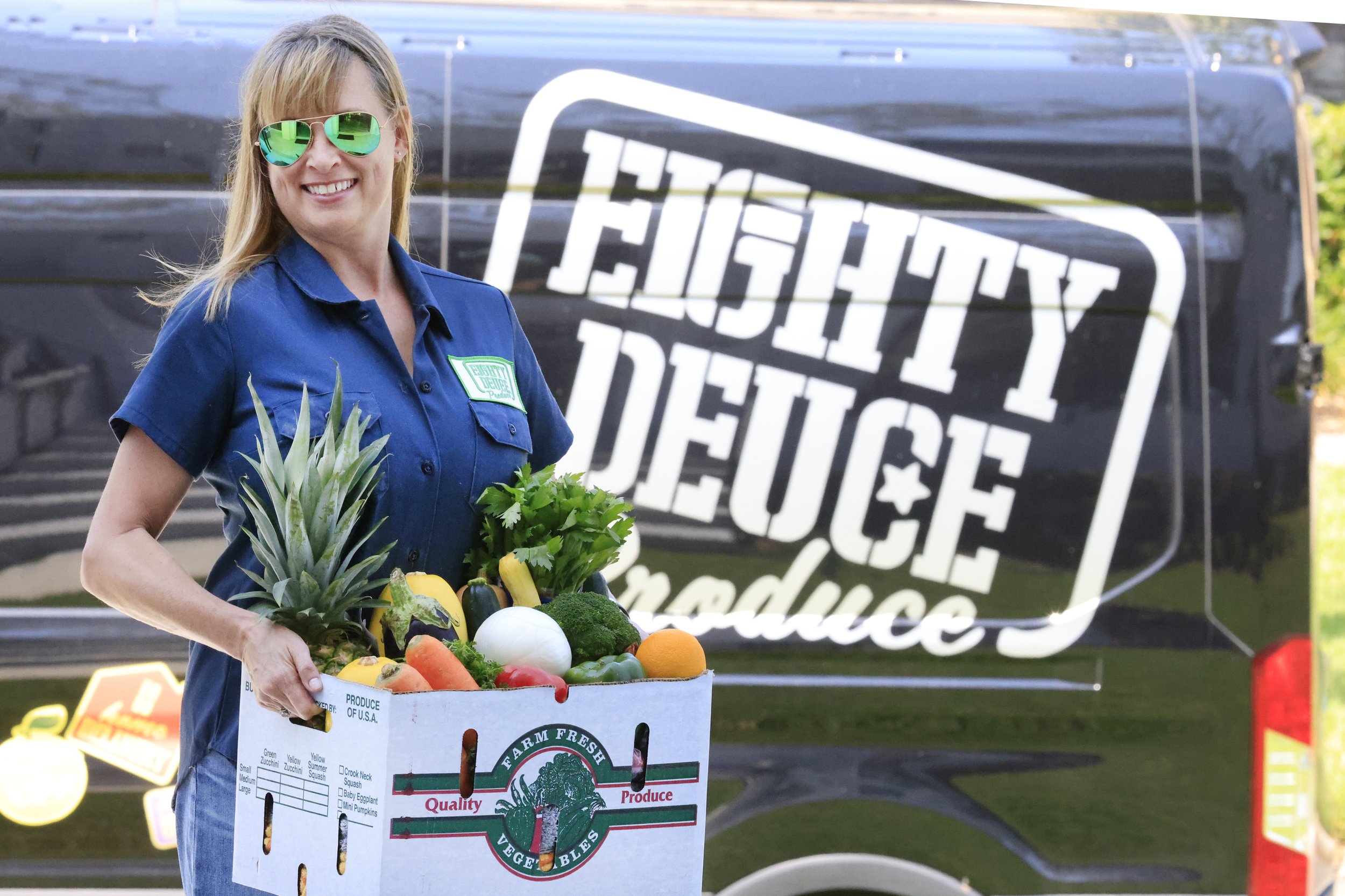 Woman wearing sunglasses smiles while holding a box filled with fresh produce, including pineapple, broccoli, and peppers, in front of a van featuring the logo of Eighty Deuce Produce.