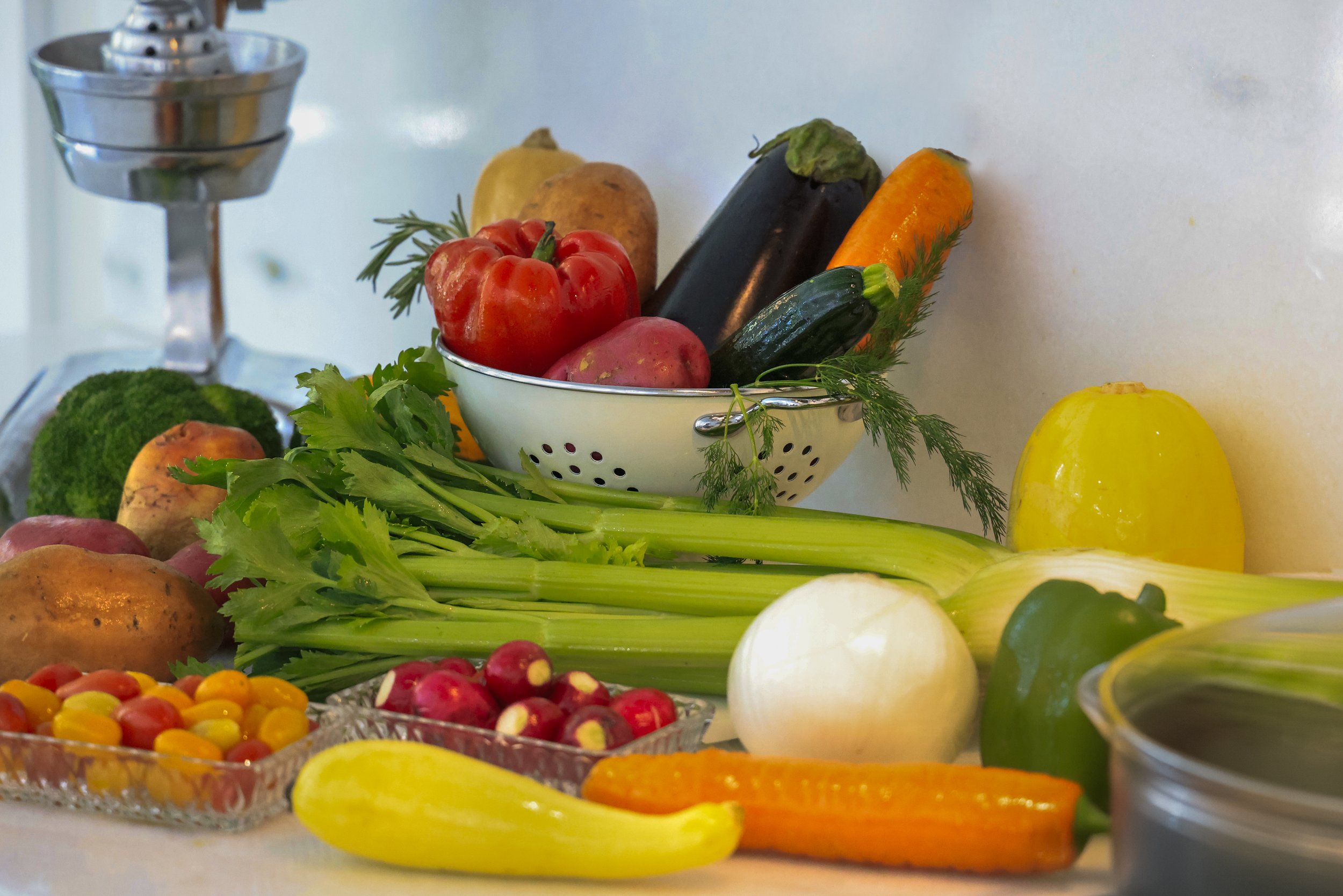 Fresh vegetables including tomatoes, zucchini, carrots, celery, and potatoes arranged on a countertop, with a colander filled with assorted produce, showcasing vibrant colors and healthy ingredients for a nutritious meal.