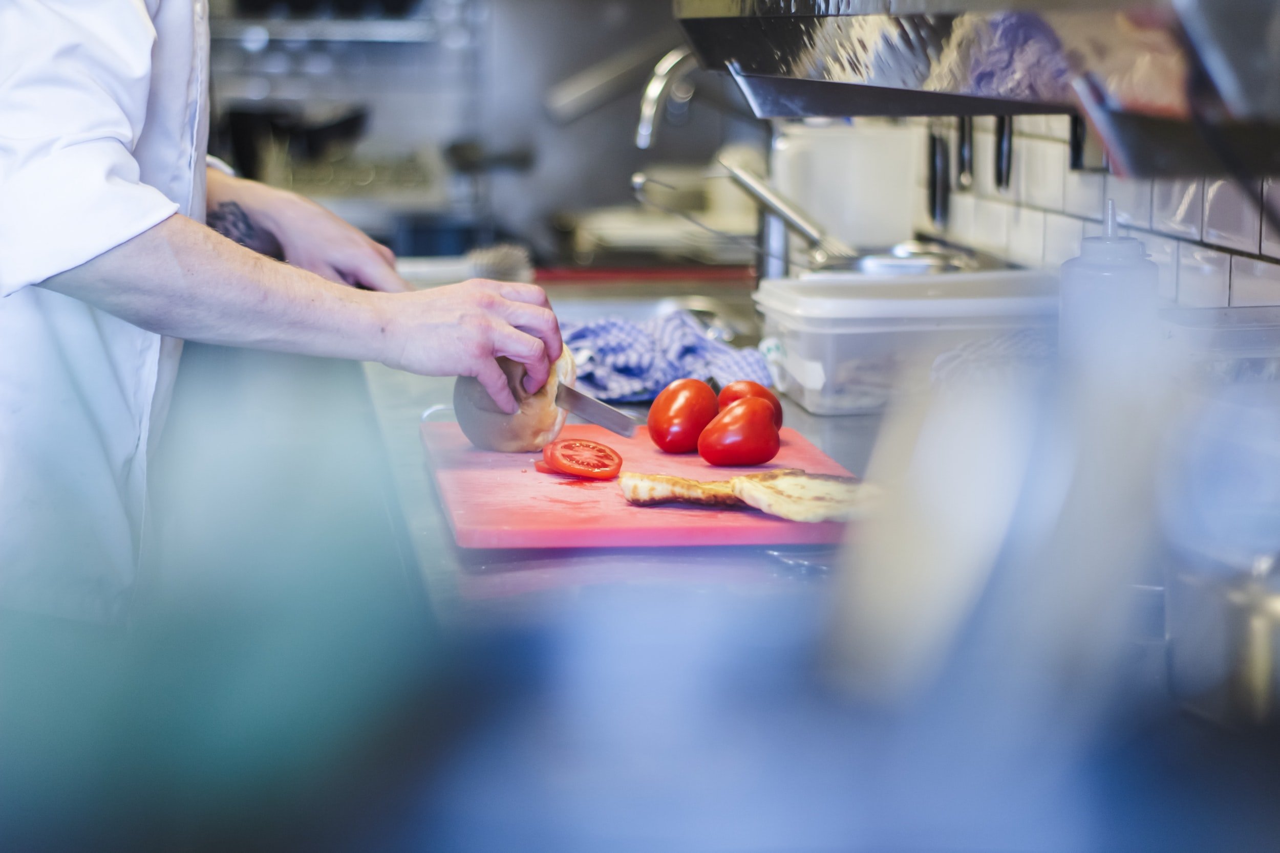 Chef preparing fresh ingredients in a professional kitchen, chopping a loaf of bread and slicing tomatoes on a red cutting board, with kitchen tools and utensils in the background.